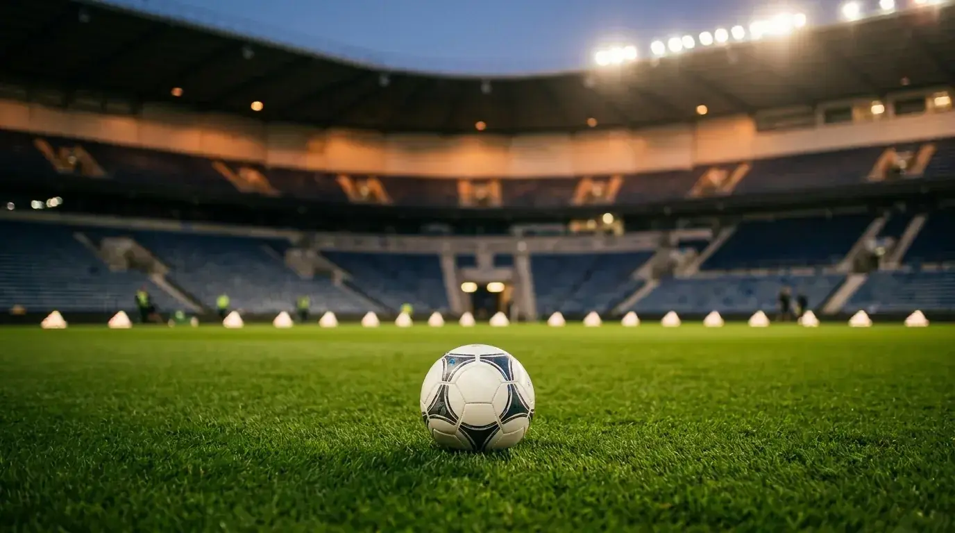 Balón de fútbol sobre el césped de un estadio iluminado al anochecer, con las gradas de fondo